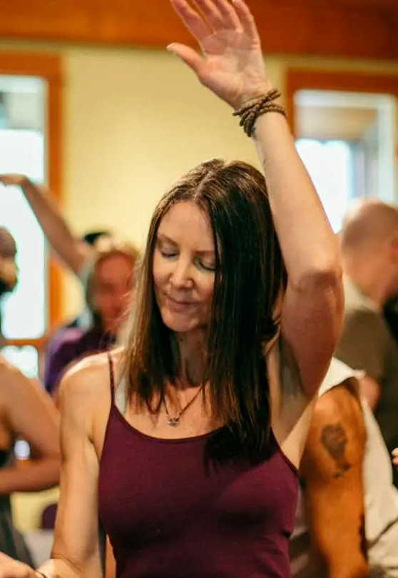 Ao fundo aparecem várias pessoas participando de uma meditação ativa no salão, na frente uma mulher com um braço para cima e olhos fechados em estado de meditação profunda, No braço levantado ela usa uma pulseira de contas e no pescoço um colar.Ela usa uma camiseta de alça fina na cor vinho.