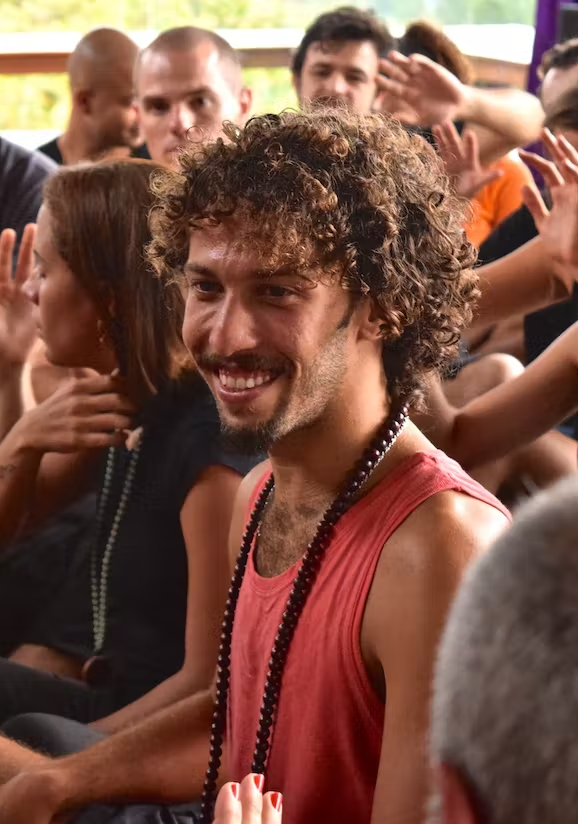Ao fundo aparecem várias pessoas em um salão participando de trabalho terapêutico, com as mãos abertas emanando boas energias para o rapaz que está no centro, ele veste uma camiseta laranjada e tem um japamala no pescoço