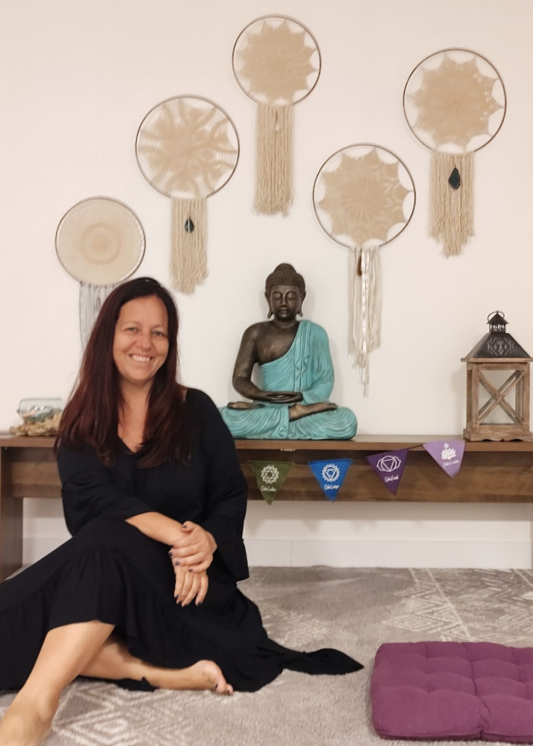 Na parede branca existem alguns filtros do sonho bege, logo abaixo uma mesa baixa de madeira com um buda com túnica azul , ao lado uma lamparina e abaixo bandeias coloridas com o símbolo dos chackras, Logo a frente a terapeuta Aline Prem Veena está sentada no chão coberto por um tapete macio bege e cinza.. Ela está com um vestido preto.